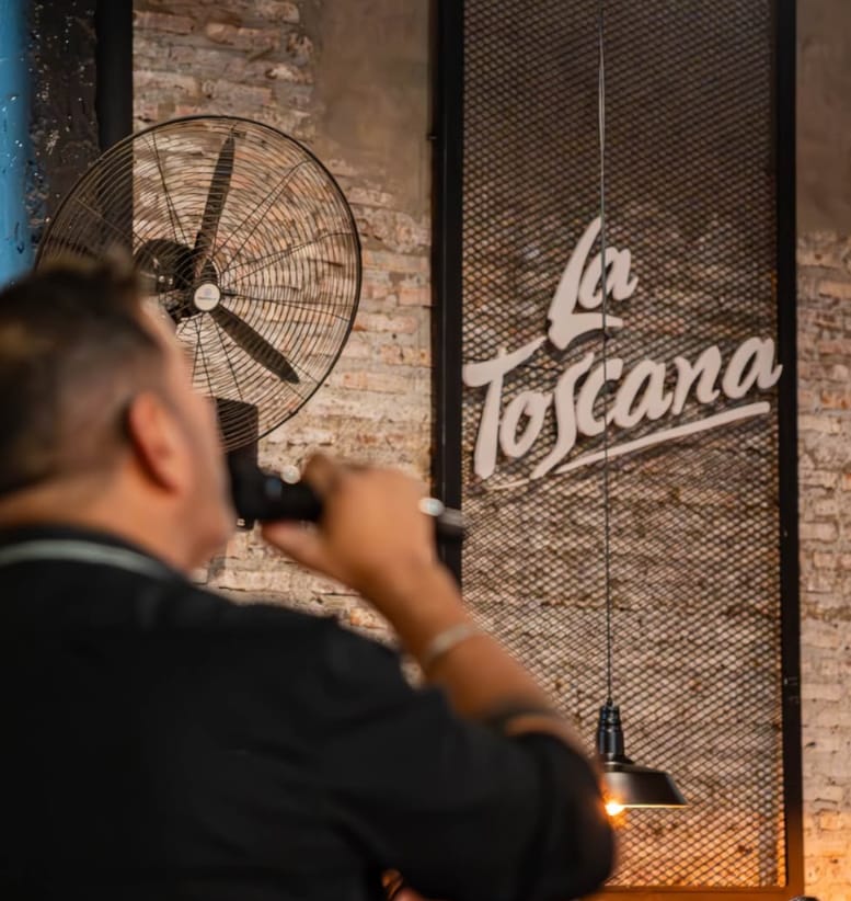 Salón de La Toscana con paredes de ladrillo, lámpara colgante y el logo en la pared; un cantante con micrófono en primer plano