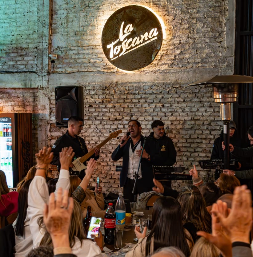 Banda tocando en vivo bajo el cartel luminoso de La Toscana mientras el público levanta las manos